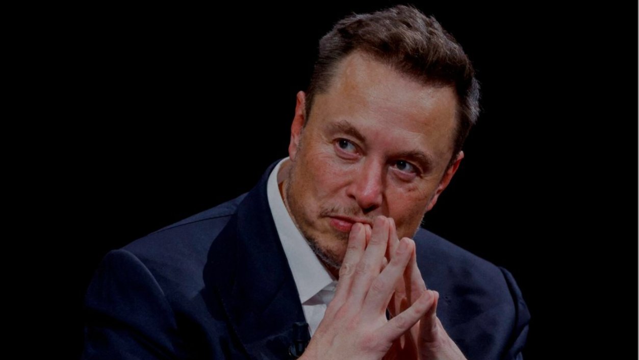 Billionaire entrepreneur Elon Musk sits in a dark suit with his hands pressed together under his chin, looking thoughtfully into the distance during an event.