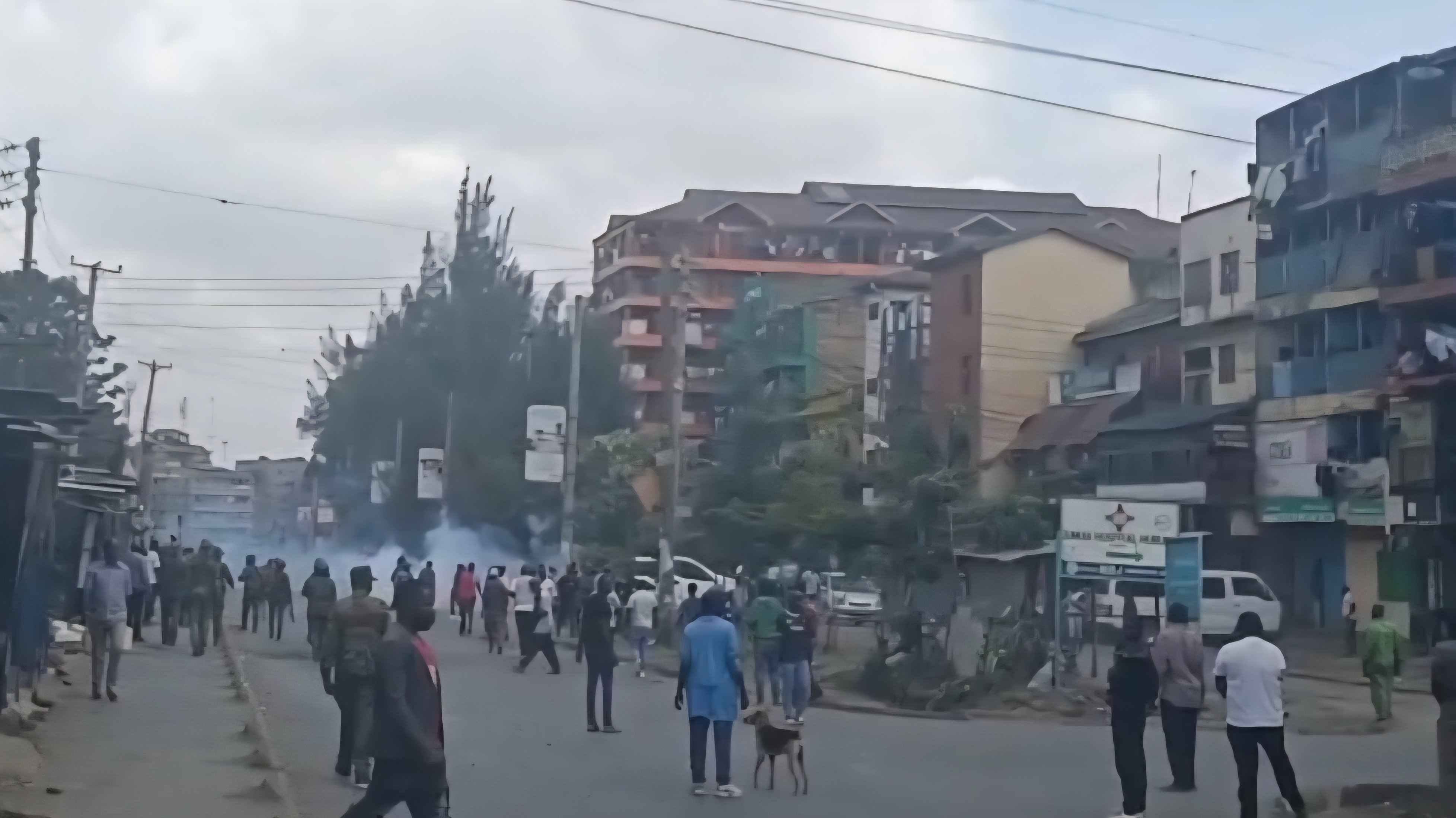 A crowd of people gathered on a paved street in Huruma with smoke in the background and residential buildings on either side.