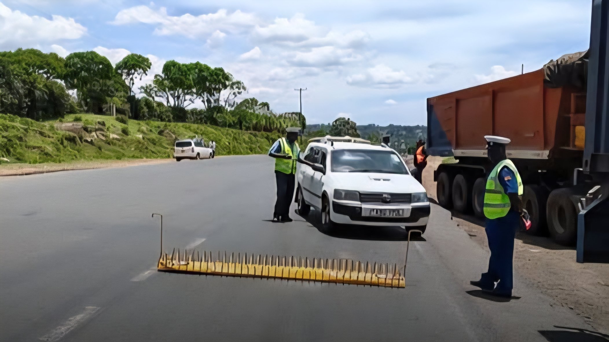 A stationary white patrol vehicle parked behind a yellow spike strip on a paved Kenyan highway, with two traffic enforcement officers inspecting the scene near a heavy transport commercial truck.