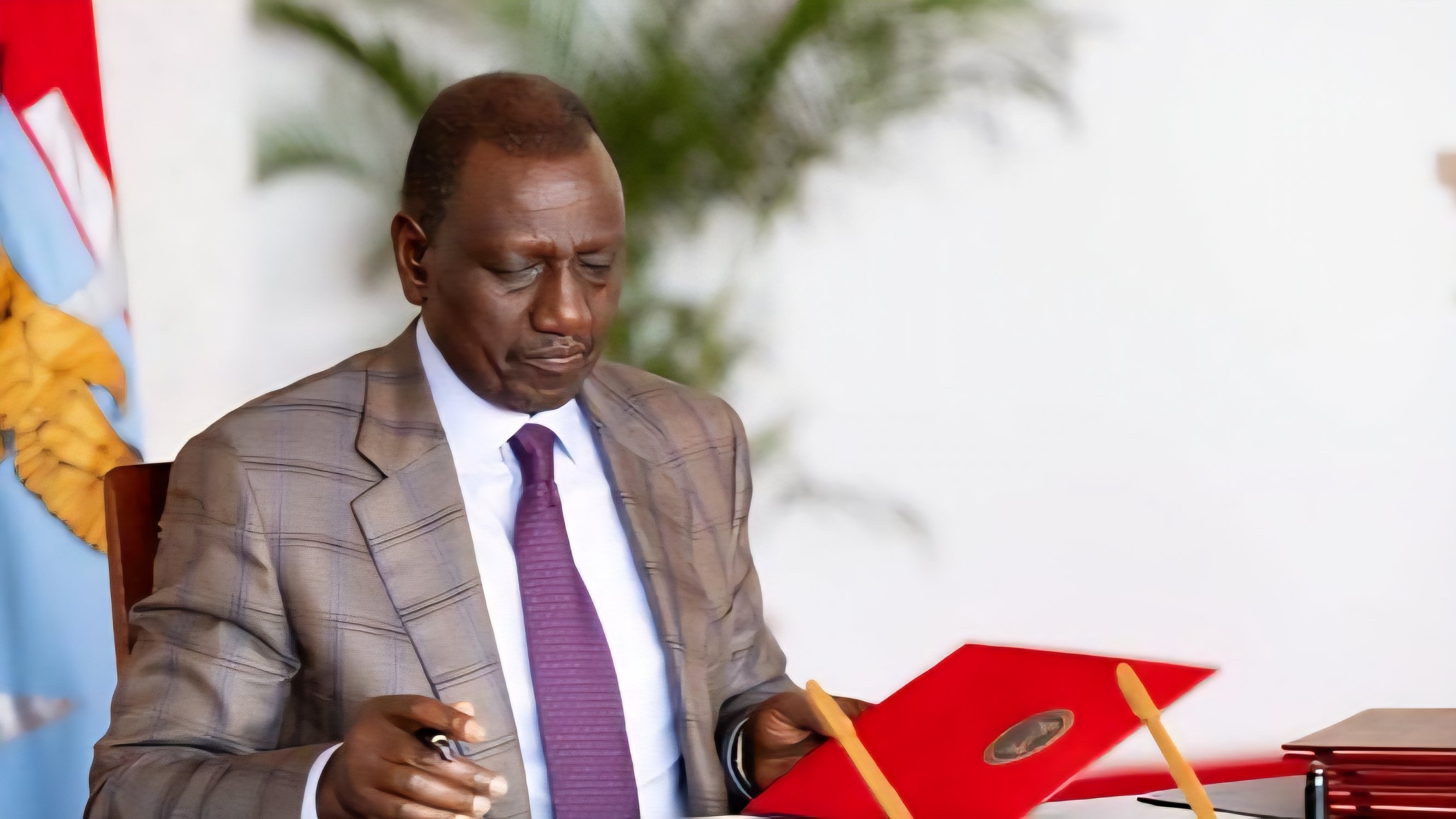 President William Ruto signing documents at a desk with a child standing beside him during an official legislative event.
