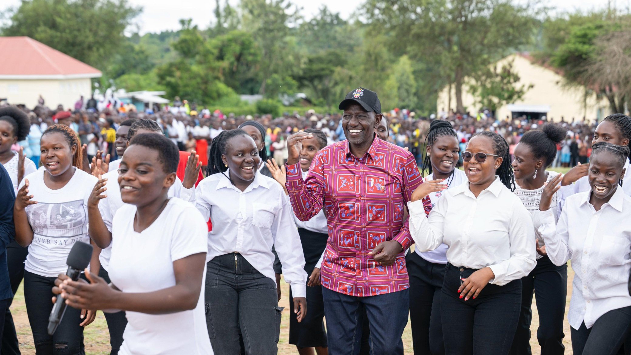 President William Ruto stands smiling among a group of young women holding a microphone during the official commissioning of Seme Teachers Training College in Kisumu County, Kenya