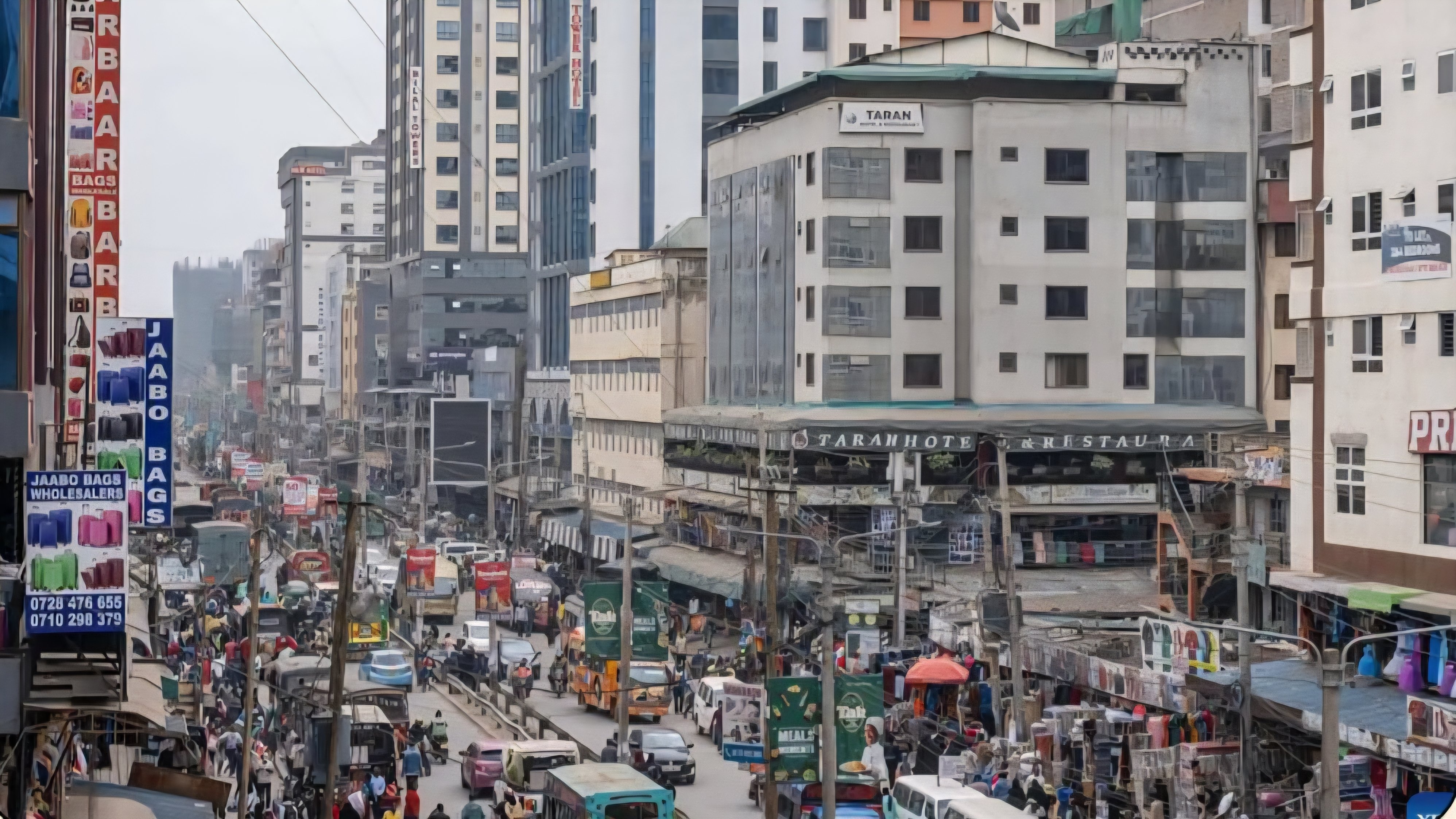 A wide-angle view of a busy commercial street in Eastleigh, Nairobi, showing multi-story trading complexes, heavy vehicle traffic, and crowded pedestrian walkways.