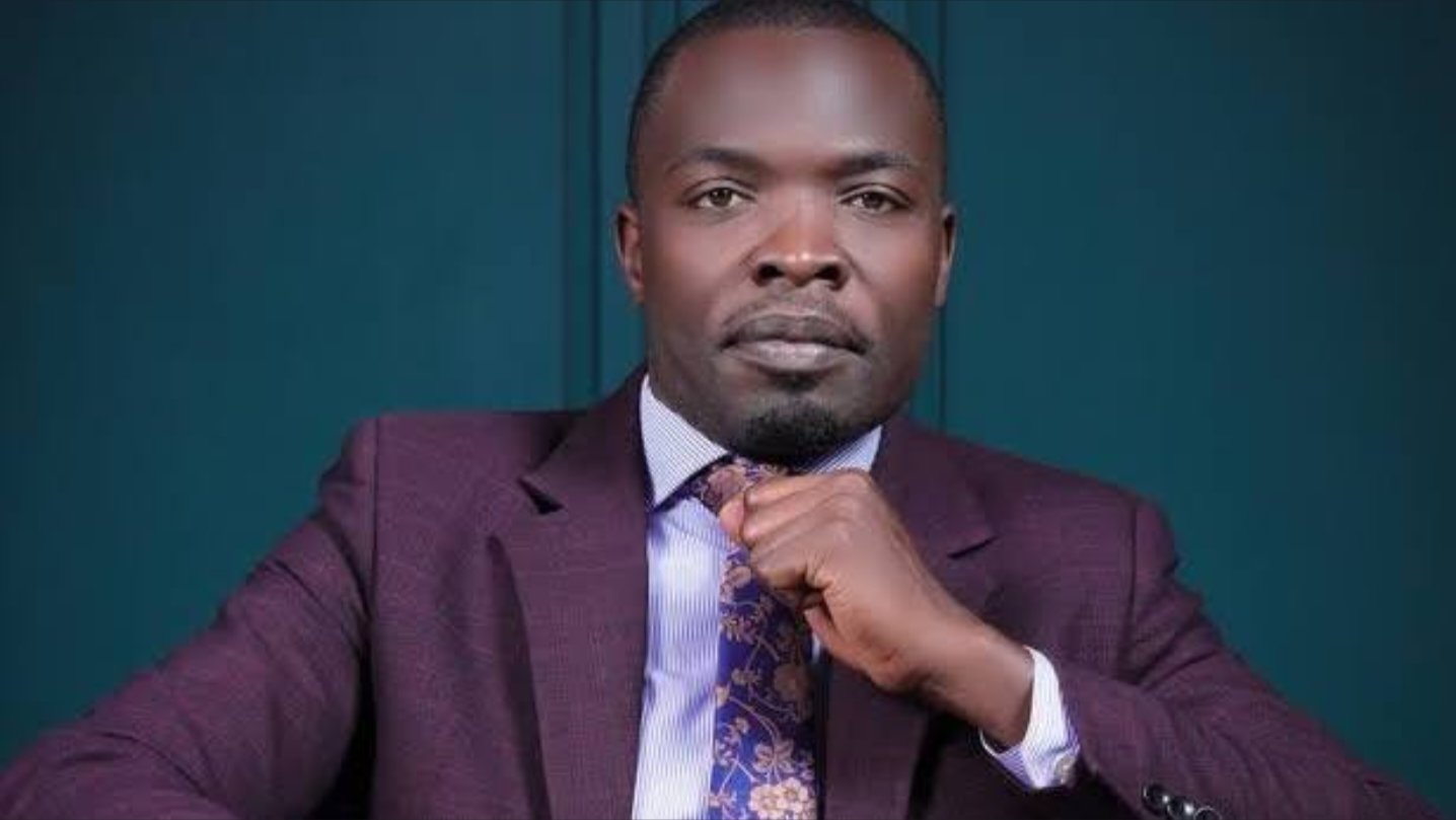A portrait of Samson Musumba, a teacher at Nairobi High School, wearing a purple suit and patterned tie while seated at a desk.