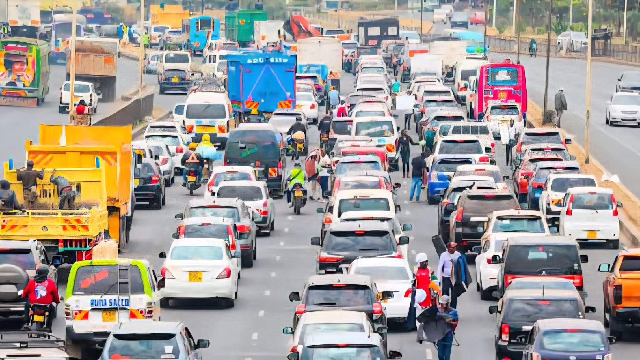 Heavy traffic congestion on a multi-lane highway in Nairobi with a retaining wall and commercial buildings in the background.