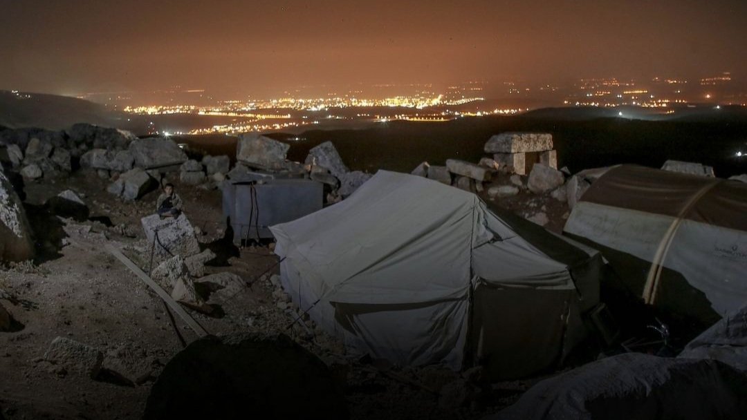 Night view of a temporary camp for internally displaced persons with white tents pitched on rocky terrain overlooking a brightly lit urban skyline in the distance.