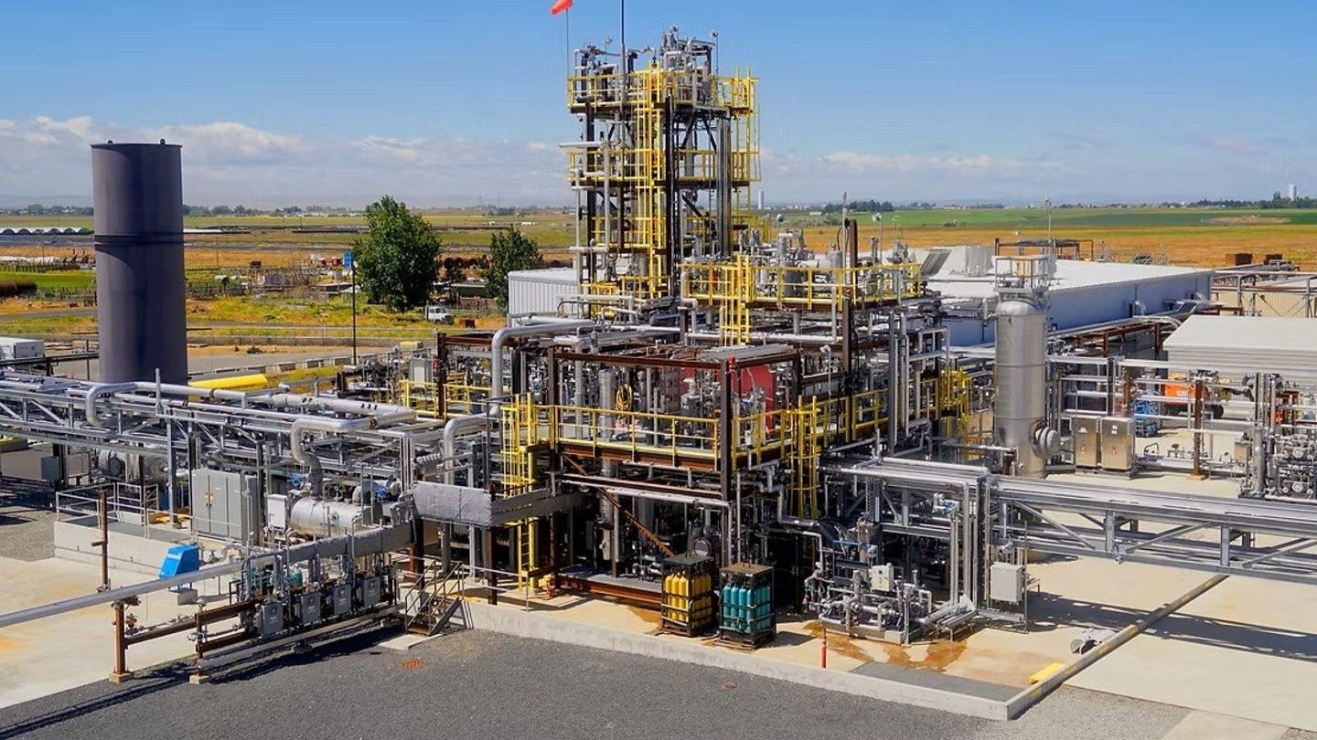 An outdoor industrial facility, AirPlant One, featuring multi-level steel scaffolding structures, complex piping, storage tanks, and yellow safety railings under a blue sky.