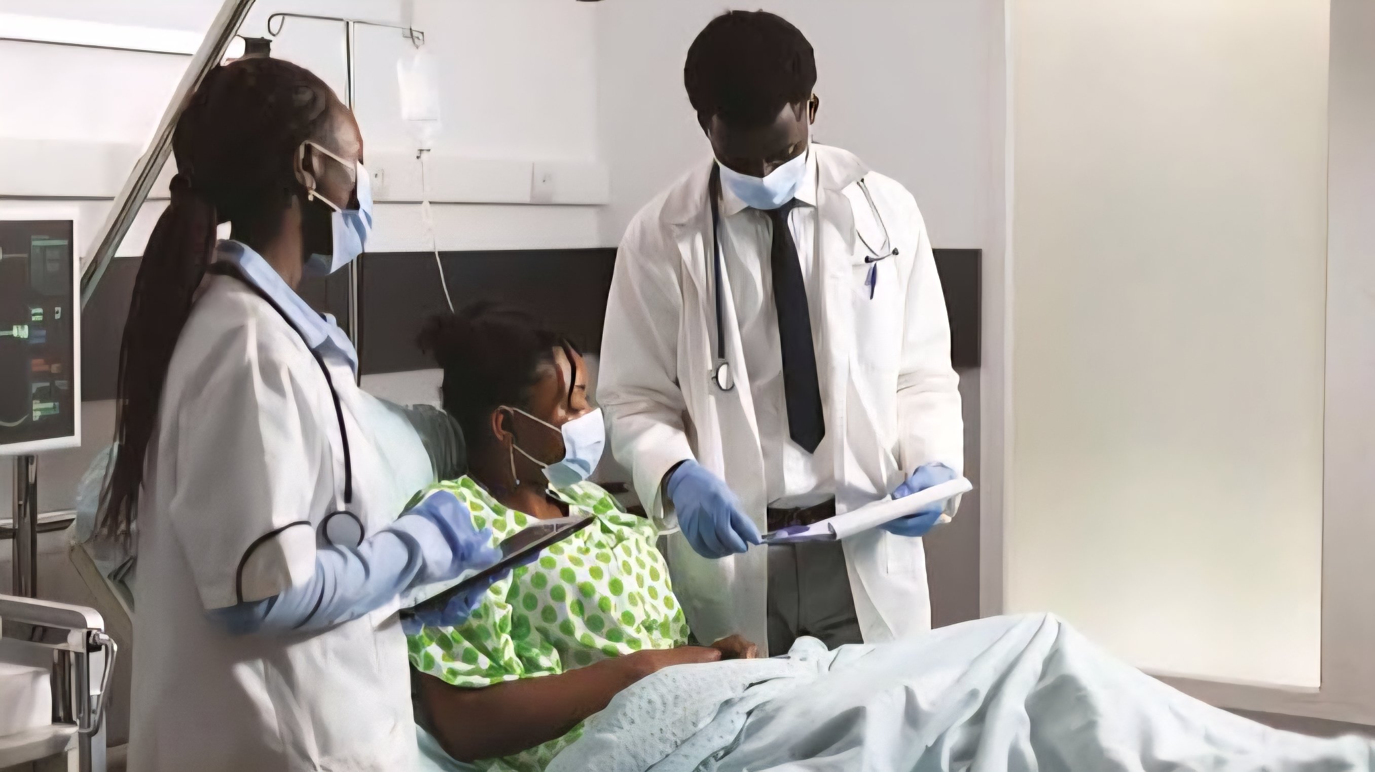 A female patient lying in a hospital bed being attended to by two medical professionals in white coats and face masks in a clinical setting.