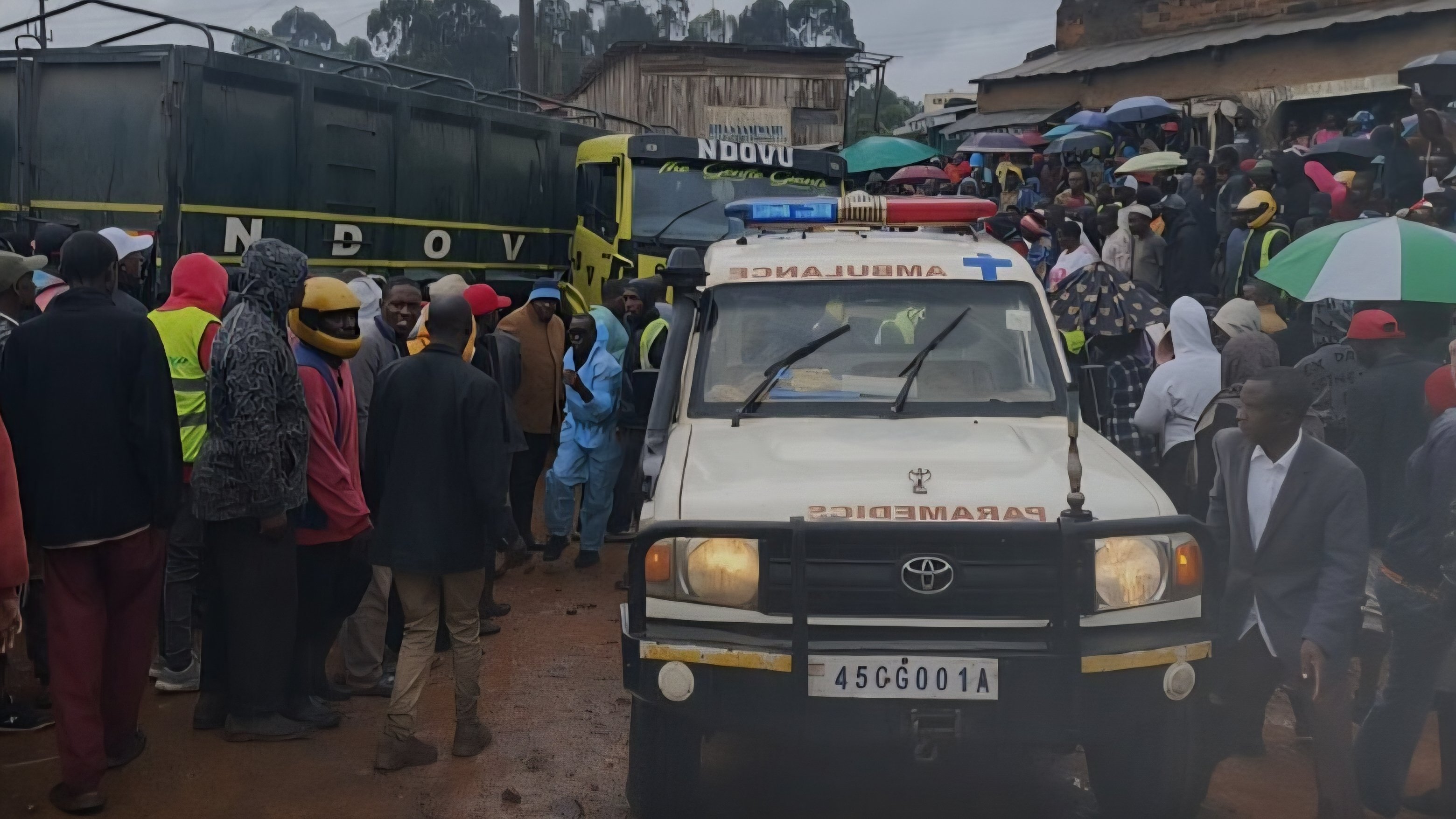 An ambulance and a crowd of people gathered at the scene of a road accident involving a trailer and boda boda motorcycles on a wet road at night.
