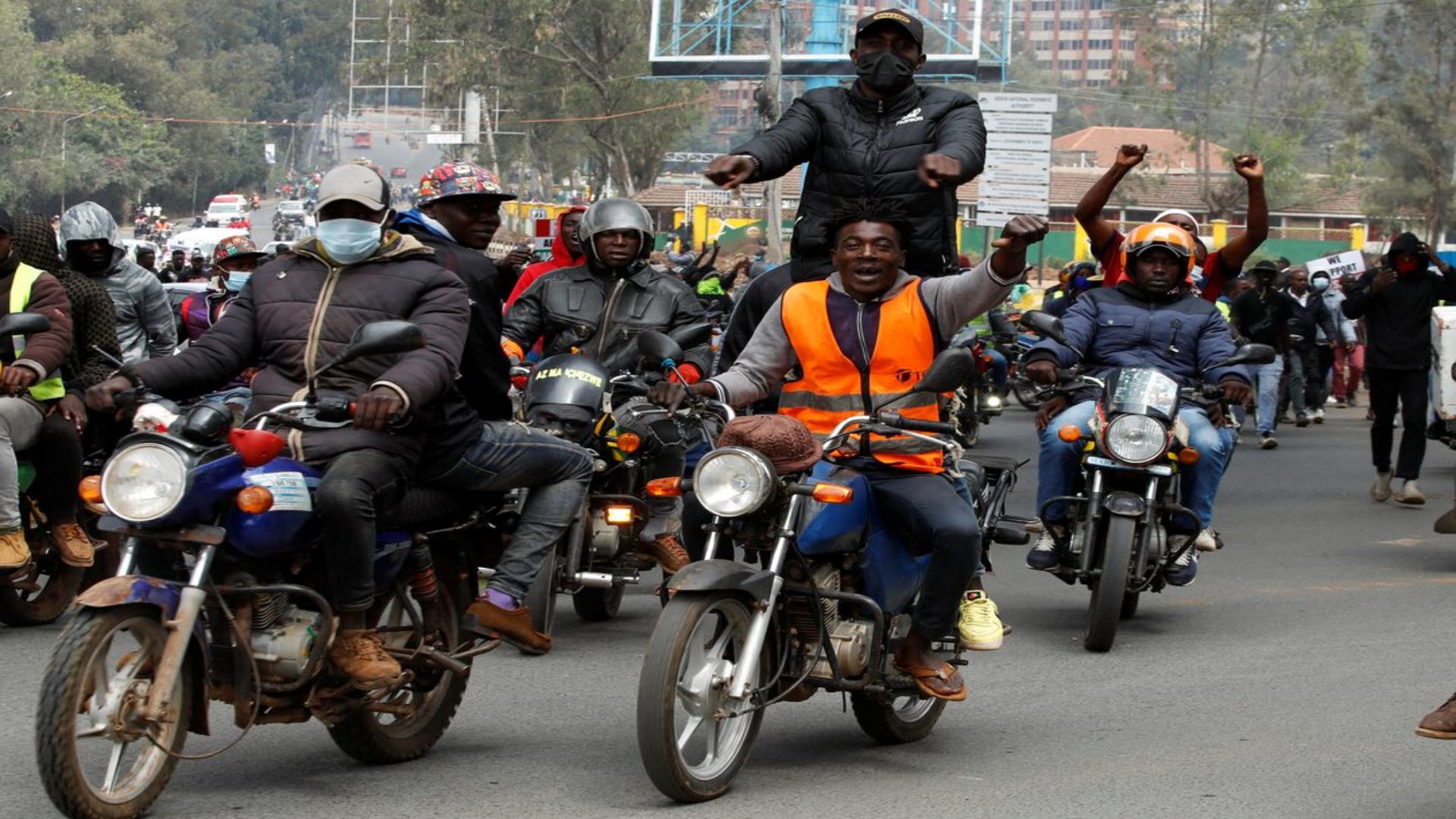 A line of motorcycles parked along a busy urban street in Kenya with riders waiting for passengers.