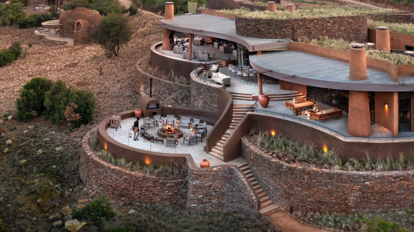 Aerial view of the contemporary stone and concrete guest area at Angama Amboseli lodge with a fire pit and sunset sky.
