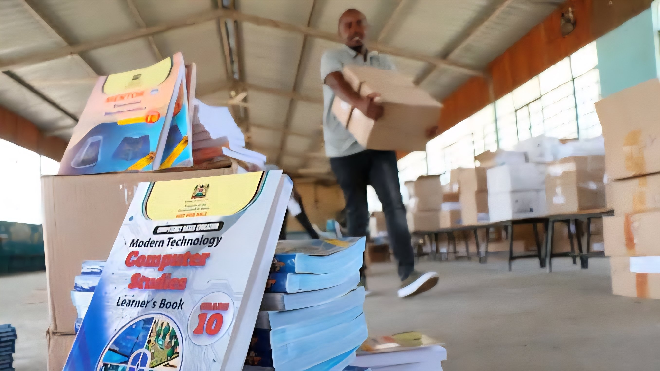 A person carrying boxes of school textbooks in a large warehouse with stacks of new curriculum books in the foreground.