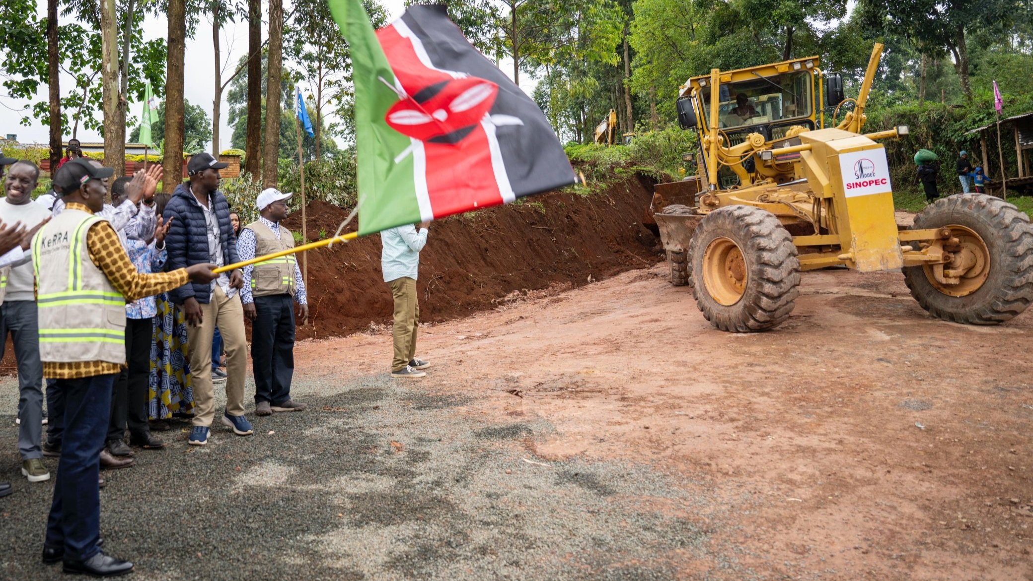 Aerial and ground-level views of a road construction launch in Kisii County, showing a yellow motor grader, a crowd of people, and Kenyan flags.