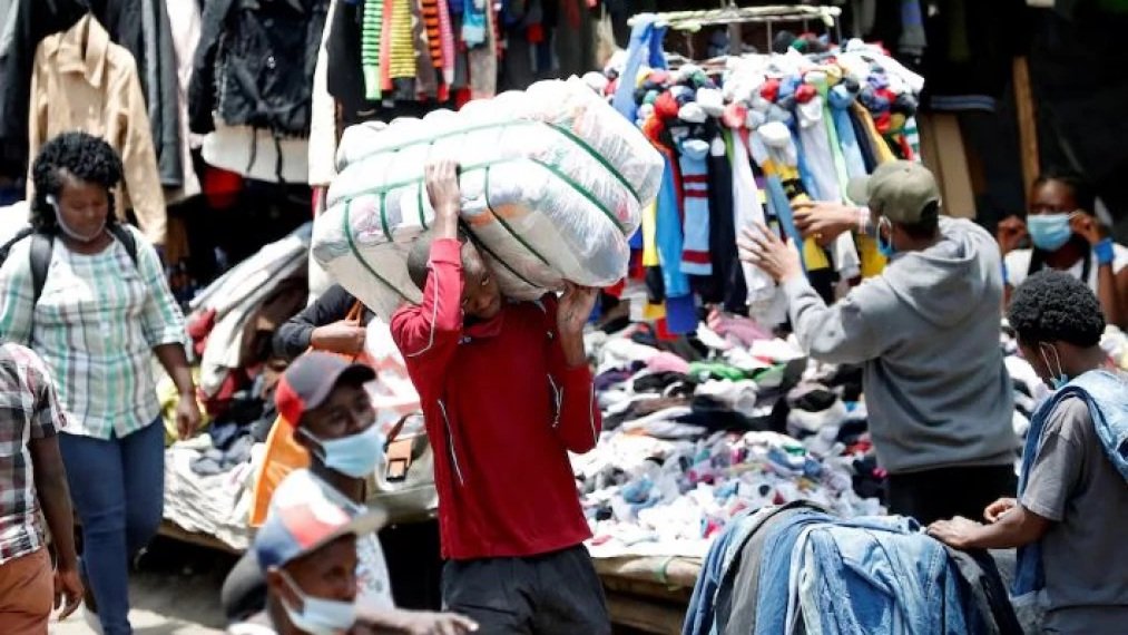 A busy day at Gikomba market in Nairobi showing a man carrying a large bale of clothes on his head through a crowded section of clothing stalls.