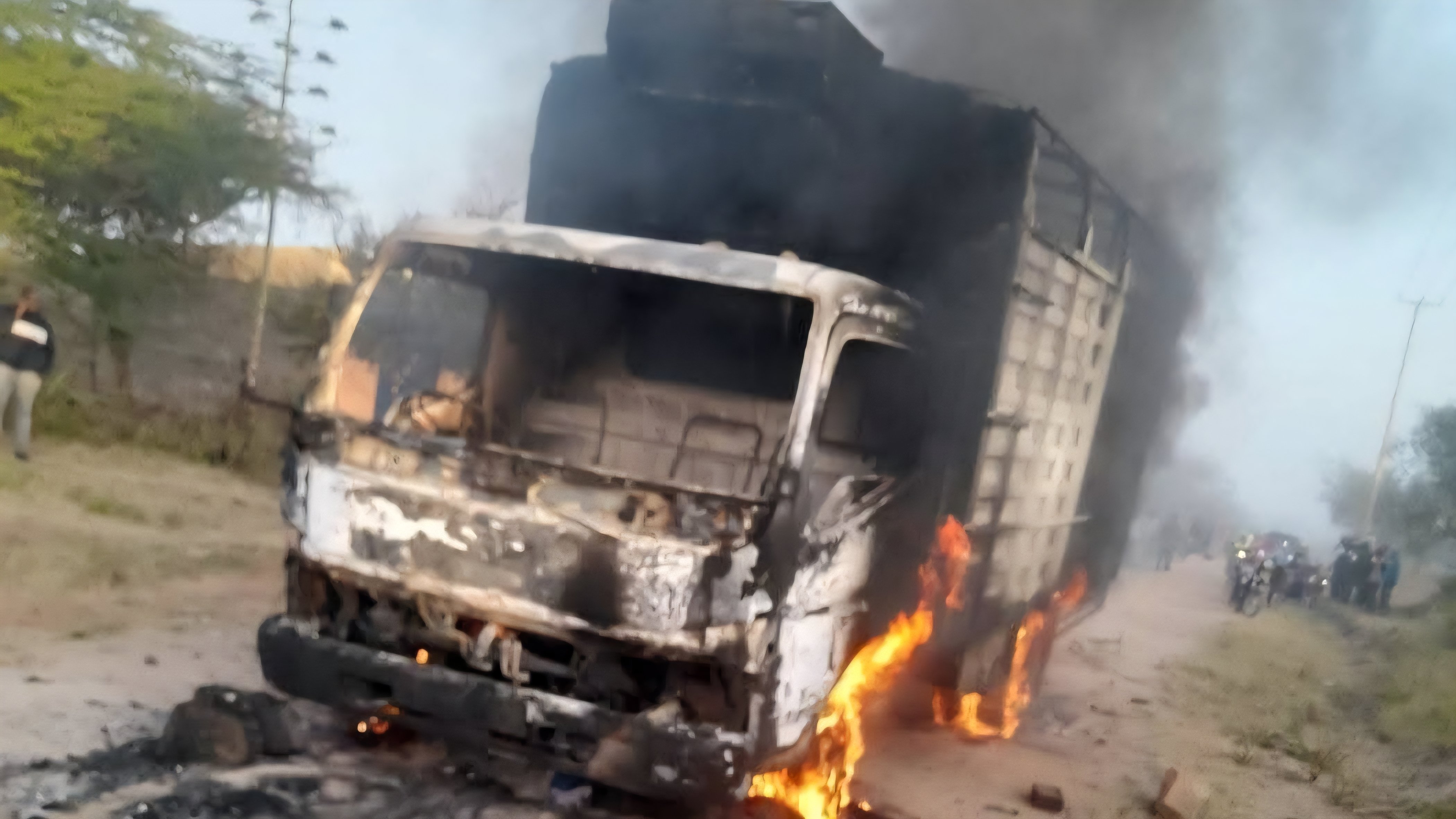 A burnt out transport lorry on a dirt road in a rural area of Makueni County with onlookers in the distance.
