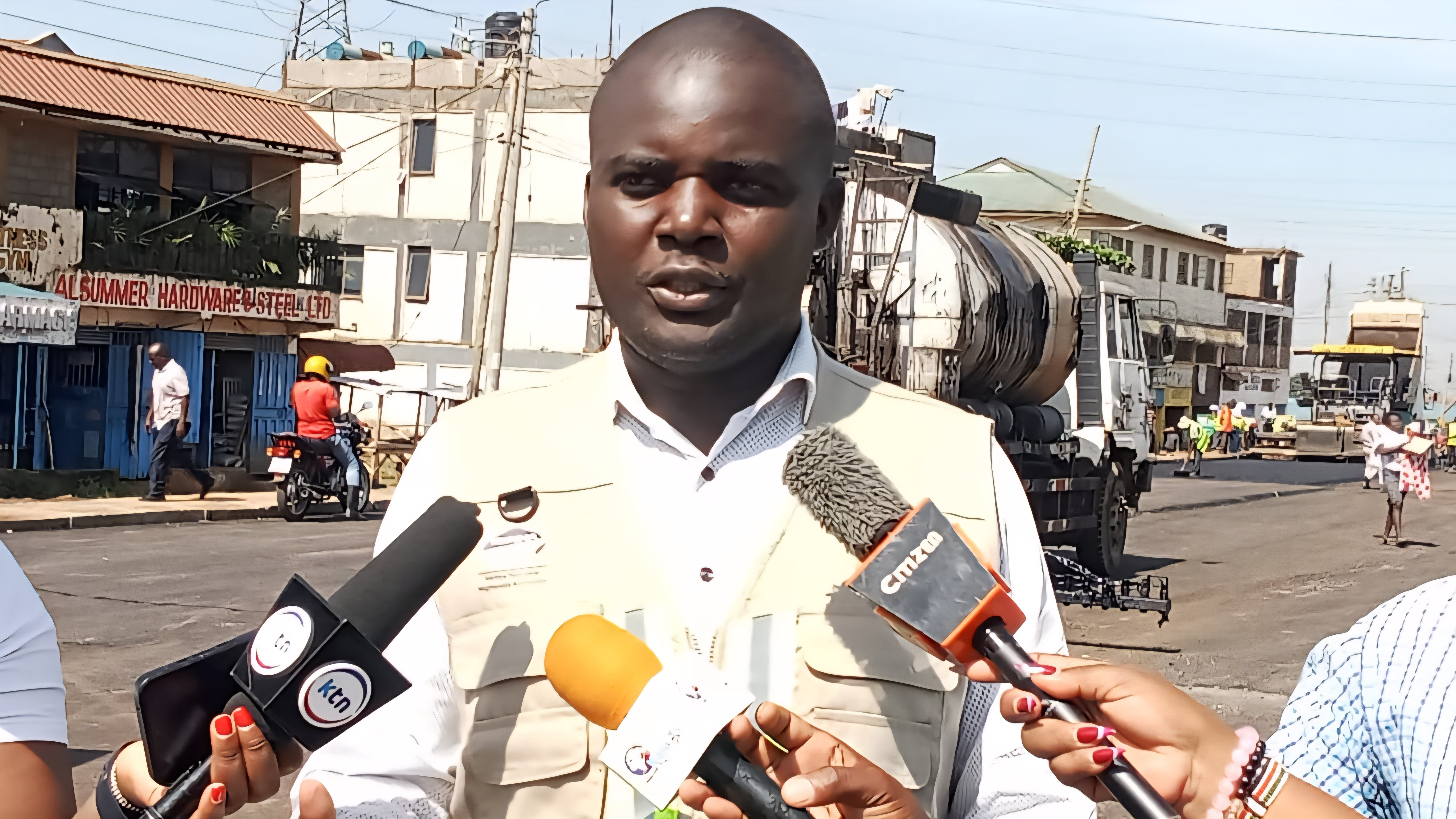 A KeNHA official addressing journalists at the Mamboleo construction site with road works visible in the background.