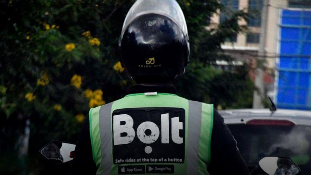 A person wearing a green Bolt branded reflector vest and a black helmet, seen from behind, sitting on a motorcycle in a city setting.