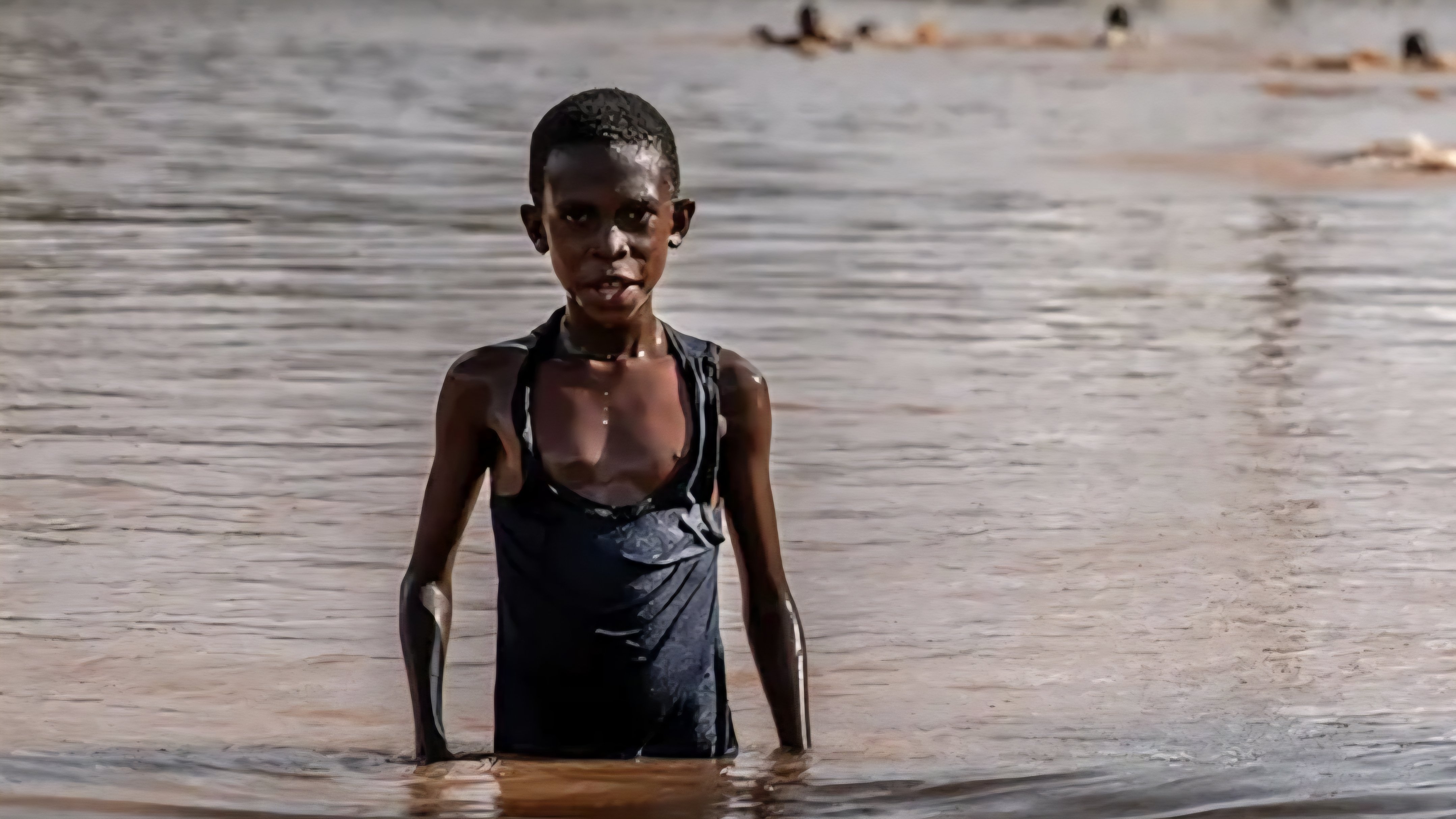 A young boy stands waist-deep in floodwaters in front of a partially submerged single-story building during a severe weather event.