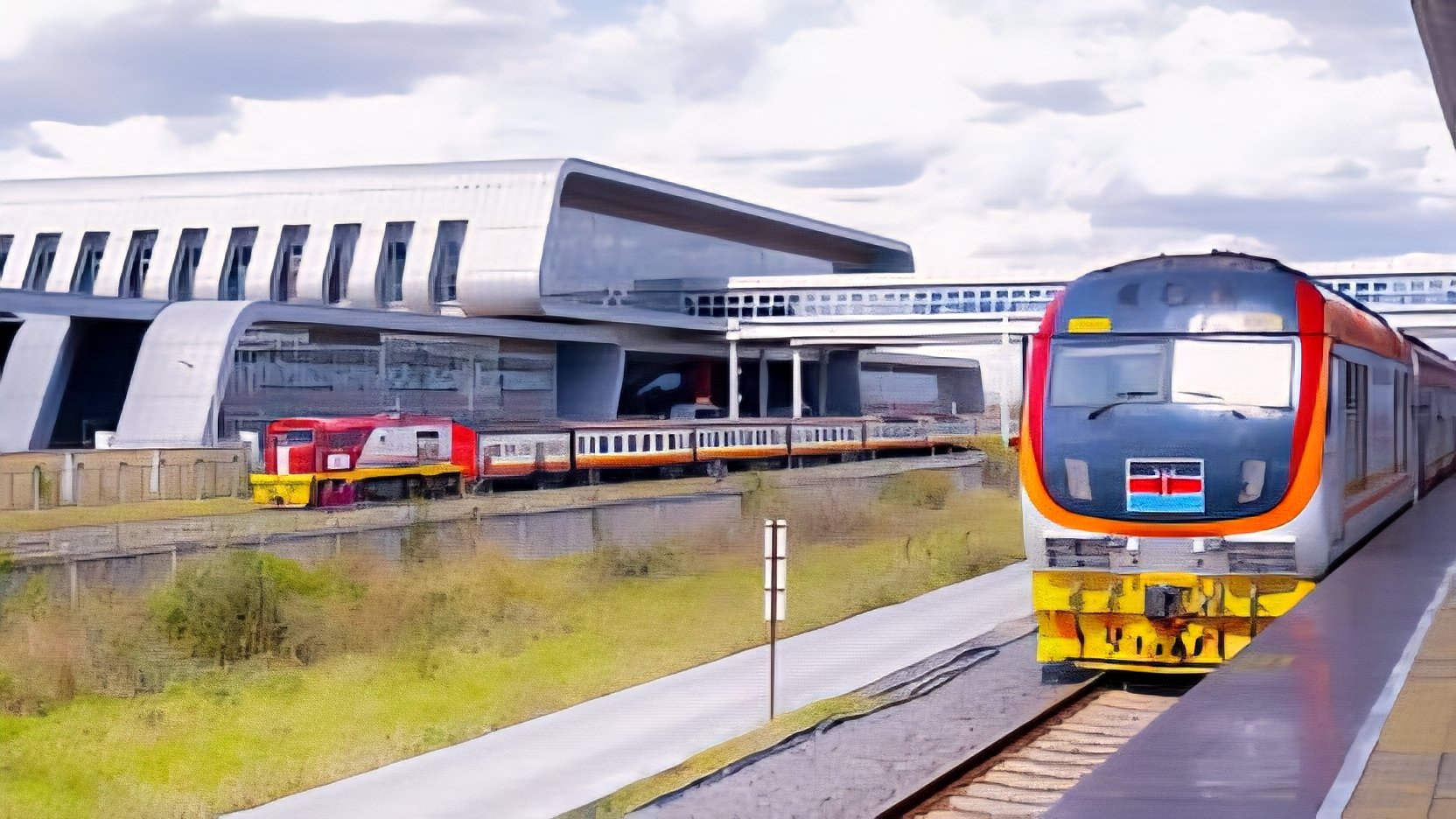 A view of a modern Kenya Railways commuter train at a station platform with the Nairobi Central Station building in the background under a cloudy sky.