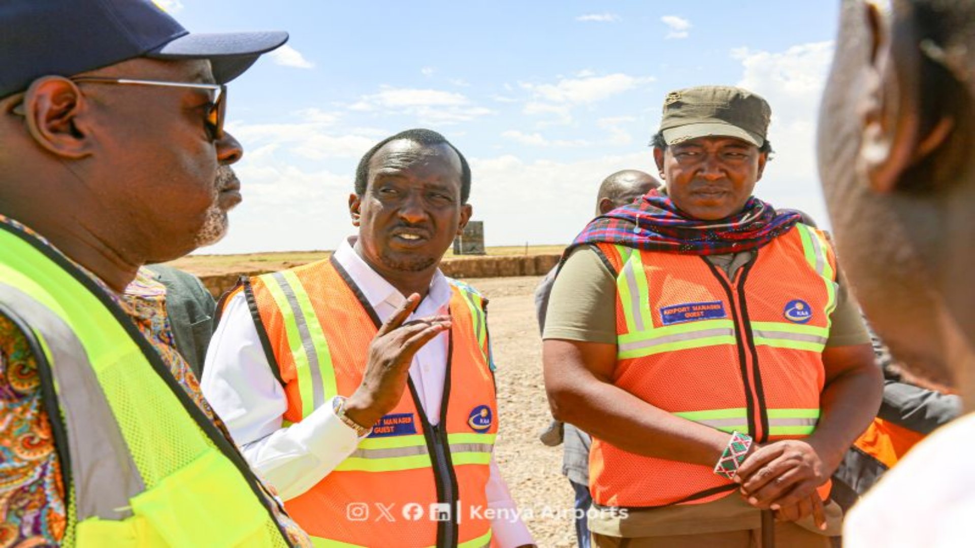Kenya Airports Authority Board Chairman Caleb Kositany shakes hands with a construction worker at the Narok Airport development site, with Narok Governor Patrick Ole Ntutu walking nearby under a clear sky.