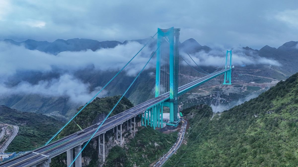 Aerial view of the Huajiang Grand Canyon Bridge in China, a massive suspension bridge spanning a deep, lush green canyon, demonstrating its extreme height and length.