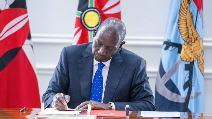 President William Ruto signing a document at a desk with the Kenyan flag in the background.