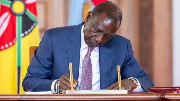 President William Ruto sitting at a formal podium during a public address with Kenyan and East African Community flags in the background.