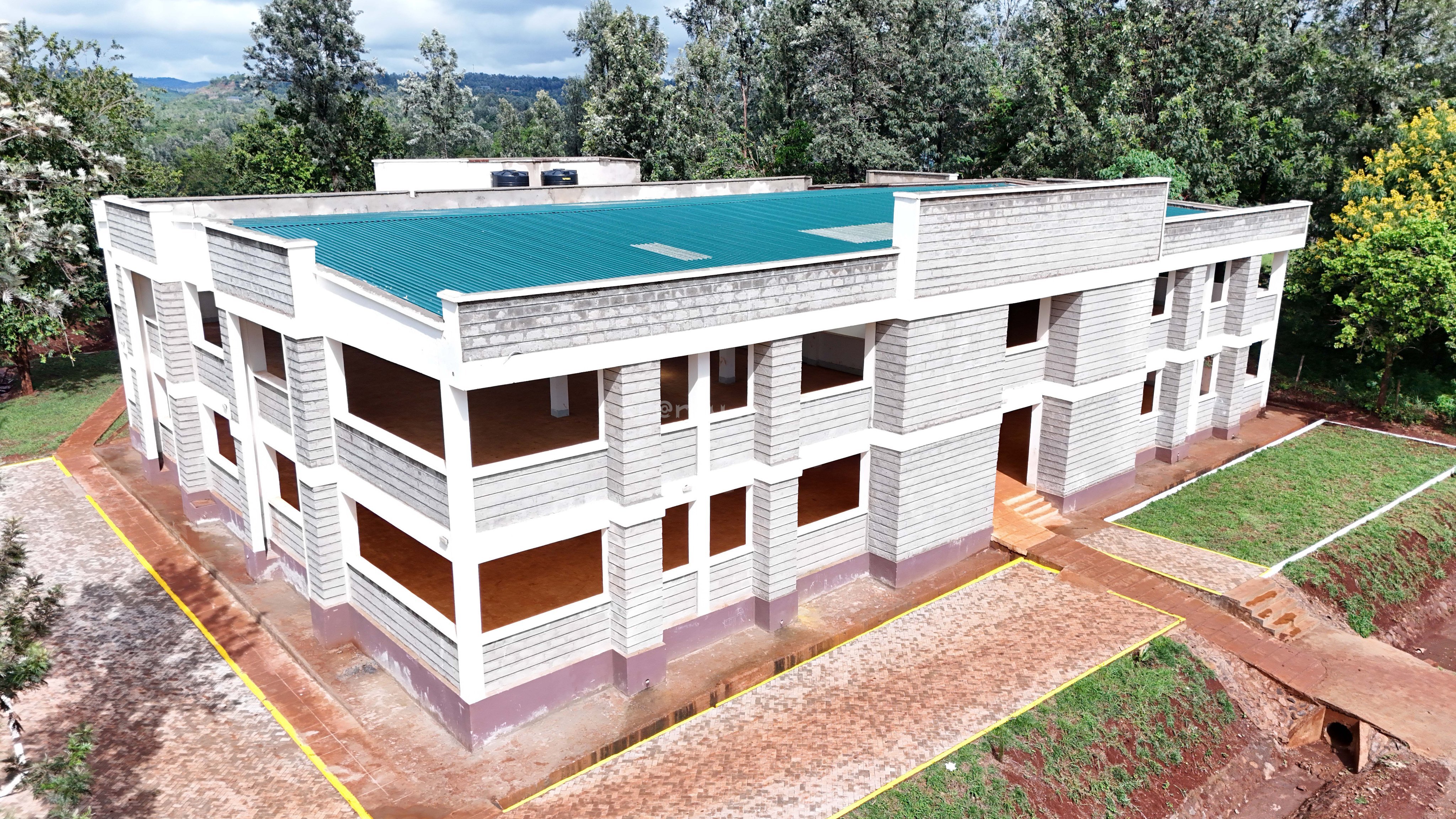 An aerial and ground-level view of the nearly completed white and maroon two-storey Kambirwa ESP Market building in Murang'a, showing the main structure, water storage tanks, and surrounding greenery.