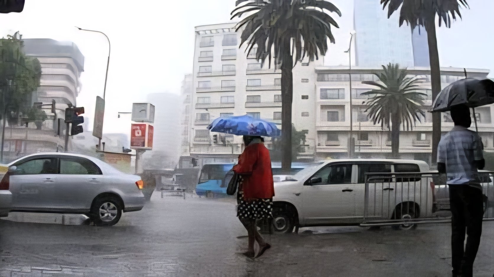 Pedestrians using umbrellas to cross a flooded street in Nairobi during a heavy downpour with vehicles and city buildings in the background.