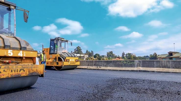 A yellow road roller compacts a fresh layer of asphalt on a new road section under construction in a rural area of Kenya.
