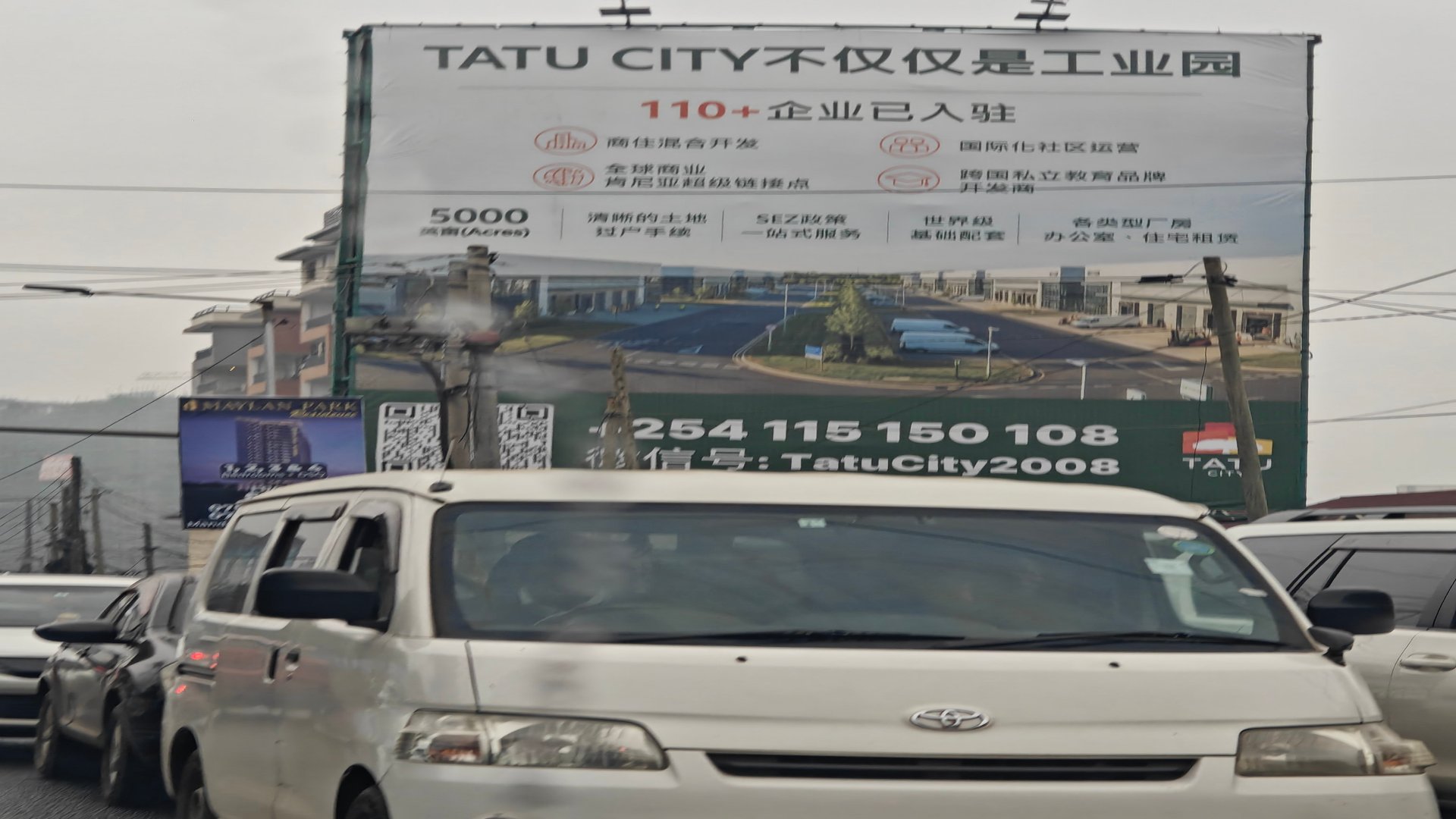 A white van driving past a large roadside billboard in Nairobi featuring Chinese text advertising real estate opportunities in Tatu City.