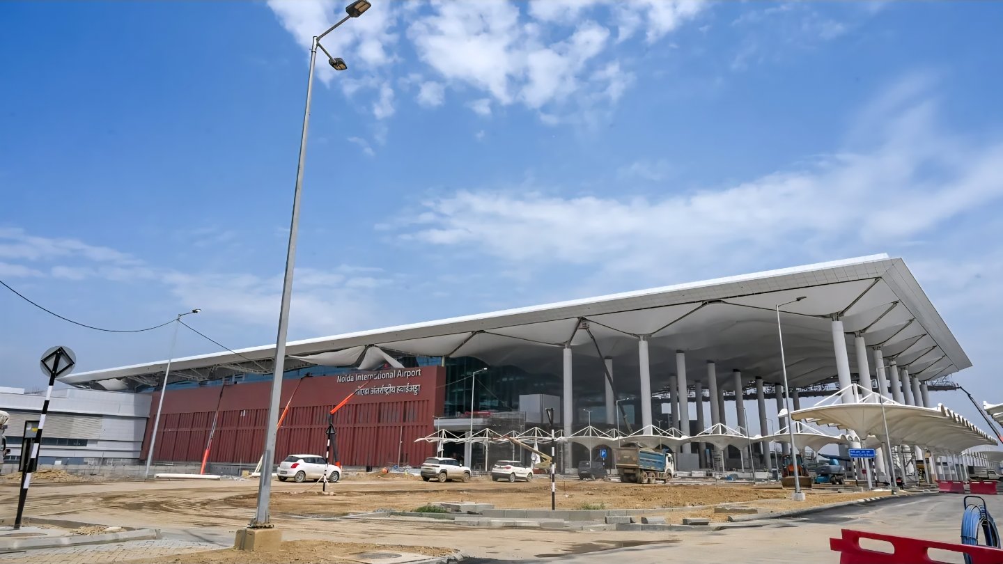 External view of the newly constructed Noida International Airport terminal building in Jewar with a large white canopy roof and support pillars under a blue sky.