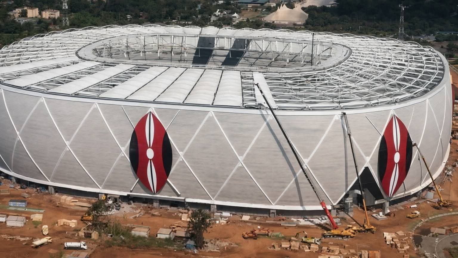Aerial view of the Talanta Sports City stadium under construction in Nairobi showing the white roof structure and large red and black shield motifs on the facade.