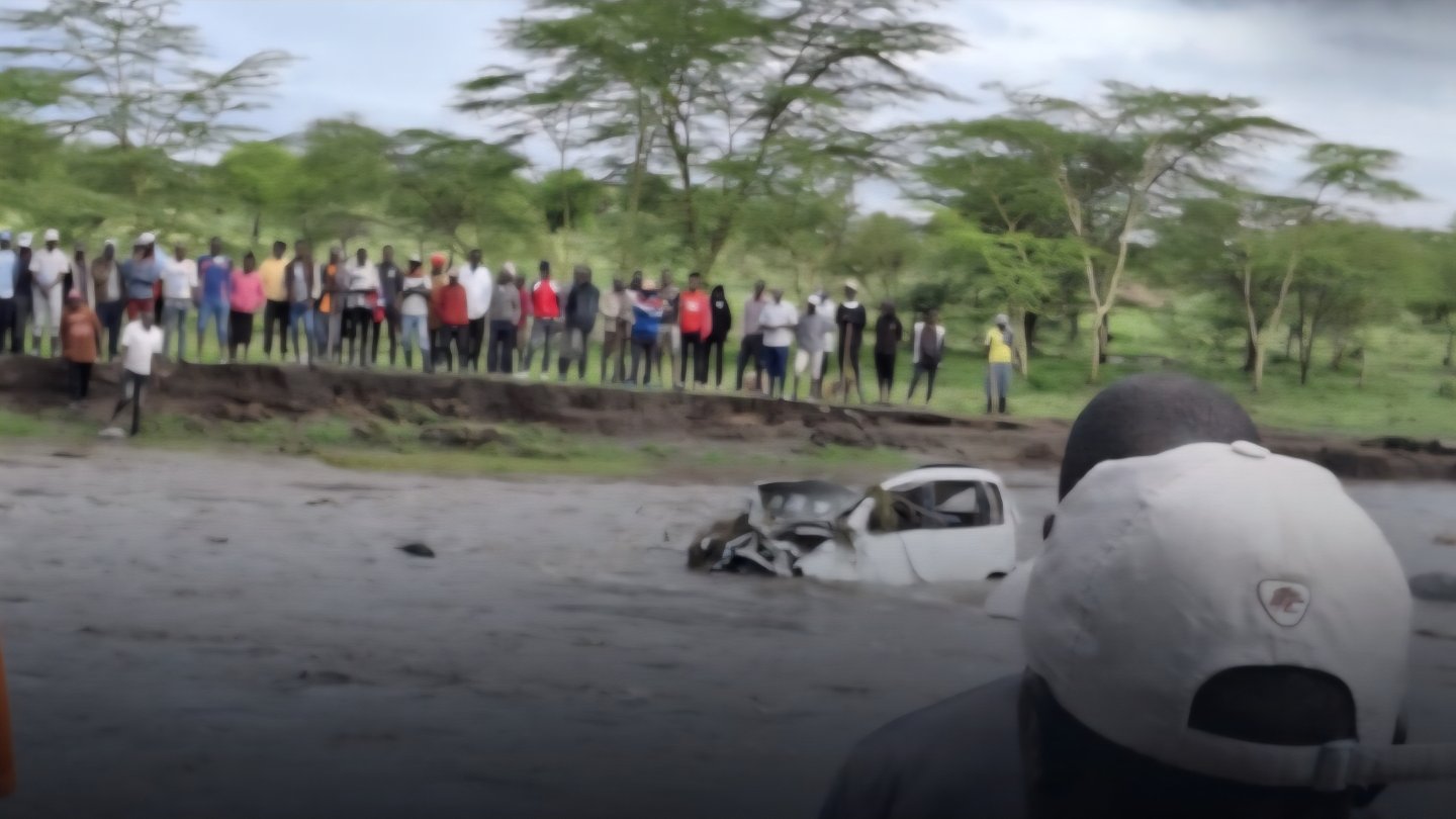A silver passenger vehicle partially submerged in a muddy floodwater channel with a crowd of people watching from a high embankment in a rural Kenyan landscape.