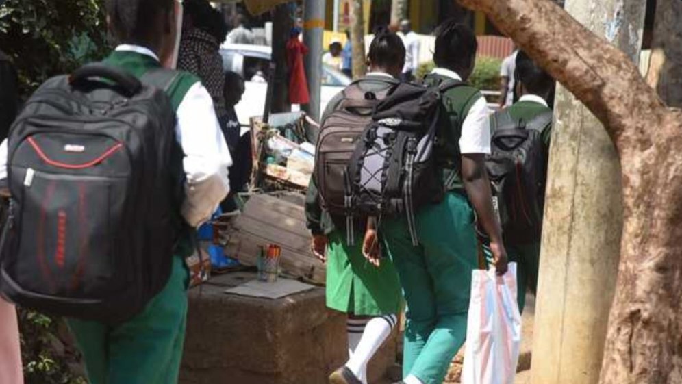 Students walking with backpacks past a concrete structure outside a Kenyan school facility.