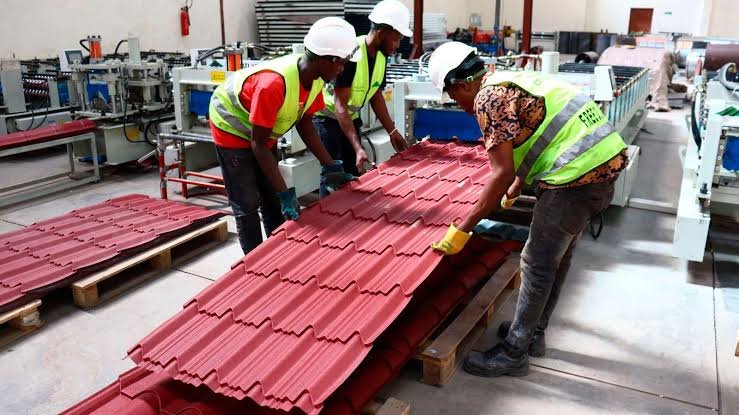 A photograph from a high angle showing industrial workers in yellow and white hard hats operating heavy machinery within a steel fabrication plant.