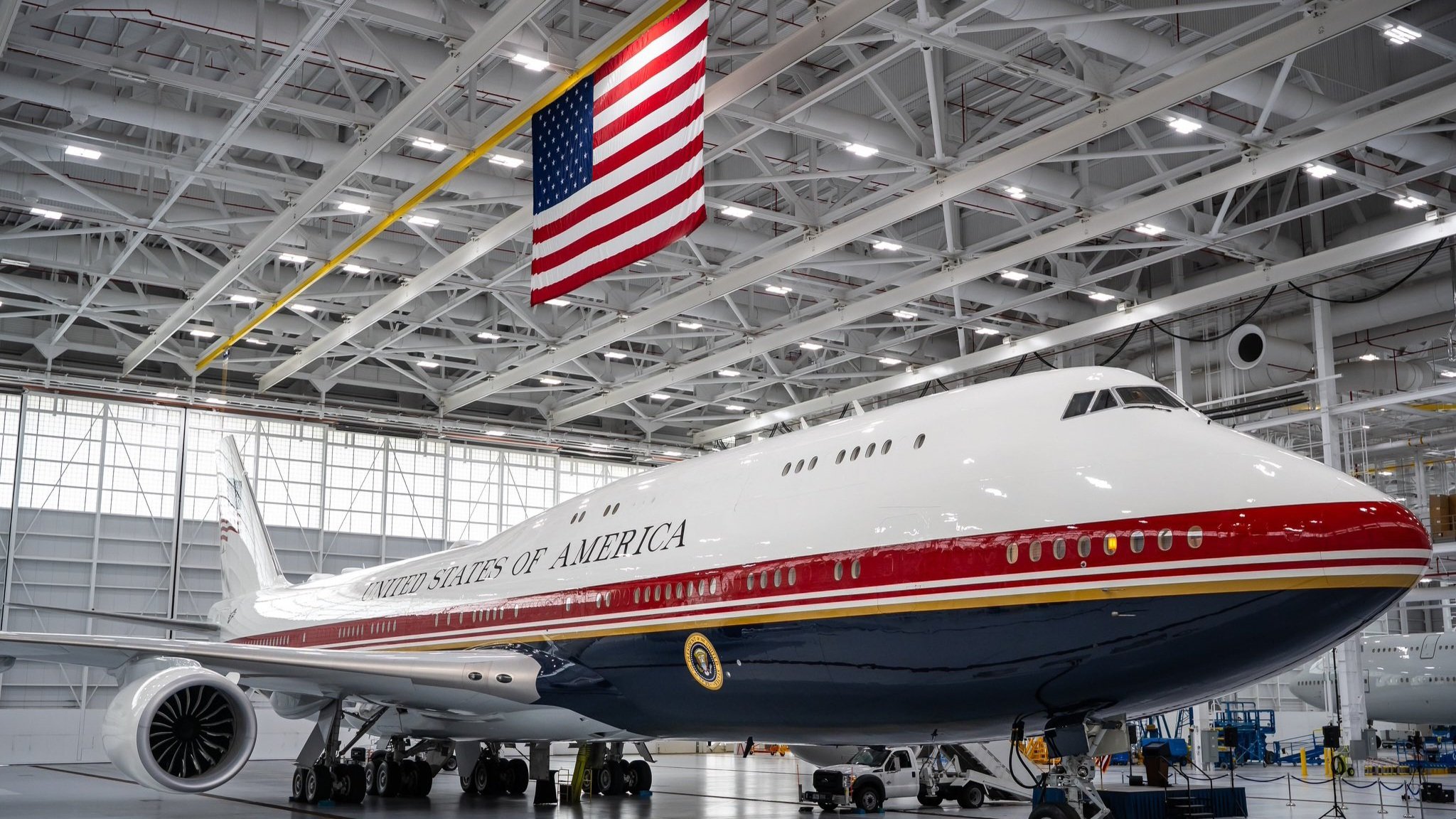 A wide interior view of a well-lit aircraft hangar showing the newly delivered VC-25B Bridge presidential aircraft, displaying its red, white, and blue livery with a prominent American flag hanging from the ceiling structure.