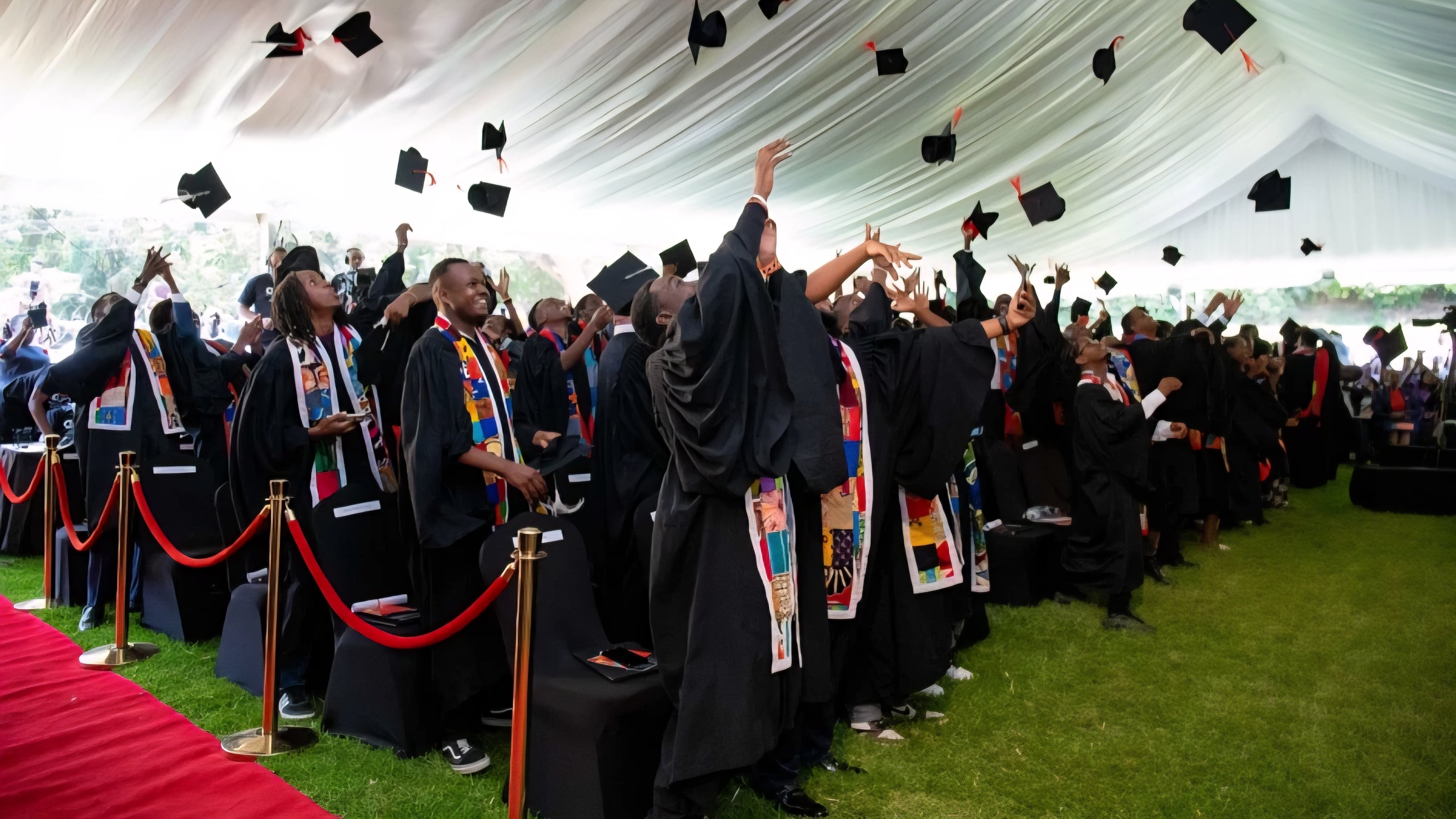 A group of graduates in traditional black academic gowns and caps standing behind a red carpet and velvet ropes during a graduation ceremony.