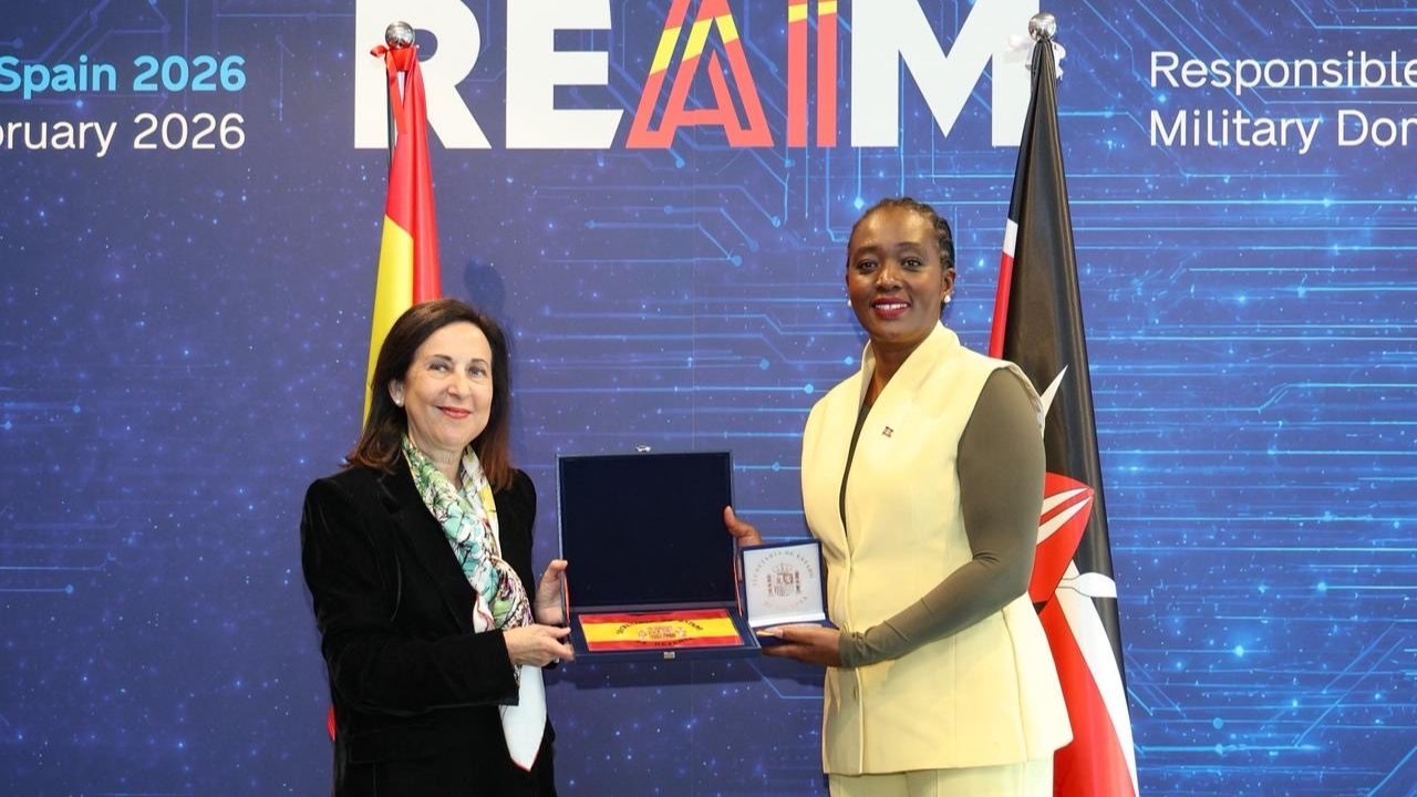 Two women exchange a plaque on stage with Spanish and Kenyan flags behind REAIM Summit backdrop.