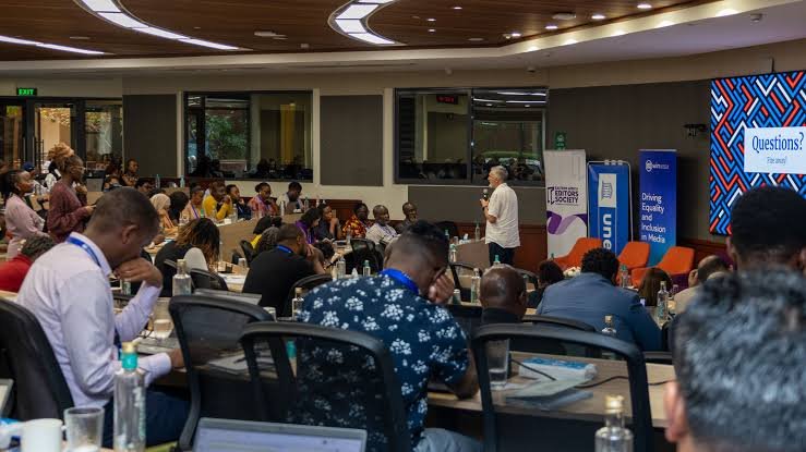 A group of media professionals and industry stakeholders standing during a conference in a modern, well-lit hall with wood-paneled ceilings.