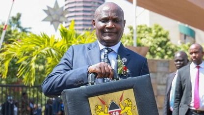 Close-up of Kenyan Treasury Cabinet Secretary John Mbadi in a formal suit, holding a black leather briefcase featuring the Kenyan Coat of Arms, during a budget-related appearance at Parliament.