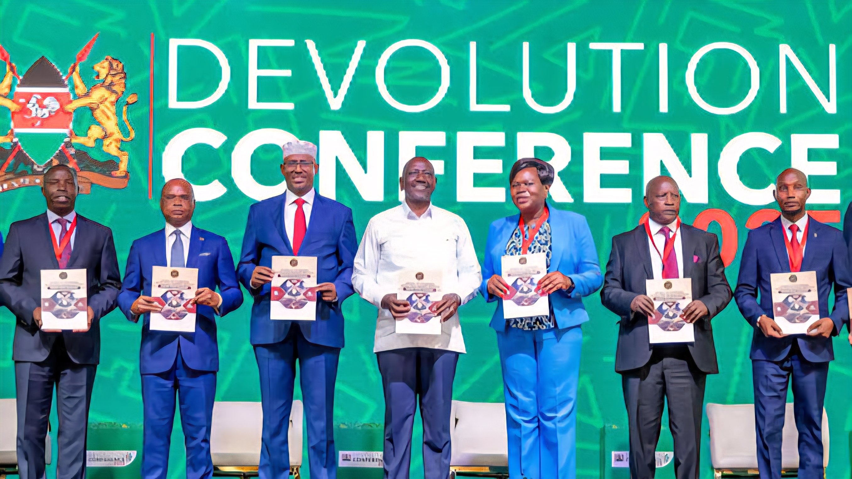 High-level Kenyan officials and dignitaries standing on a stage during the Devolution Conference, holding official documents.