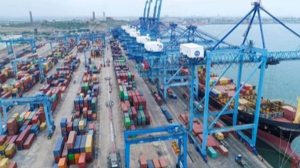 Shipping containers stacked at a busy port terminal with cranes and vessels in the background