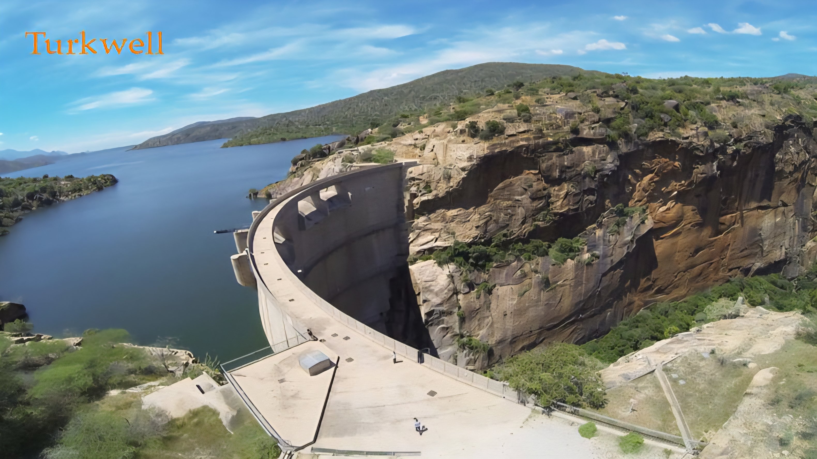 A wide aerial view showing the concrete arch wall of the Turkwel Dam in Kenya, with a deep blue reservoir on one side and the steep rocky cliffs of the gorge on the other.