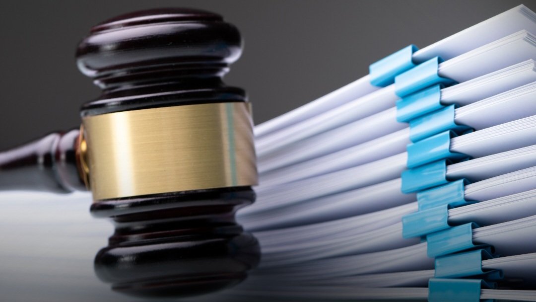 A wooden judge's gavel rests next to a large stack of legal files and corporate documents bound with blue clips, as shown in file 248955.png.