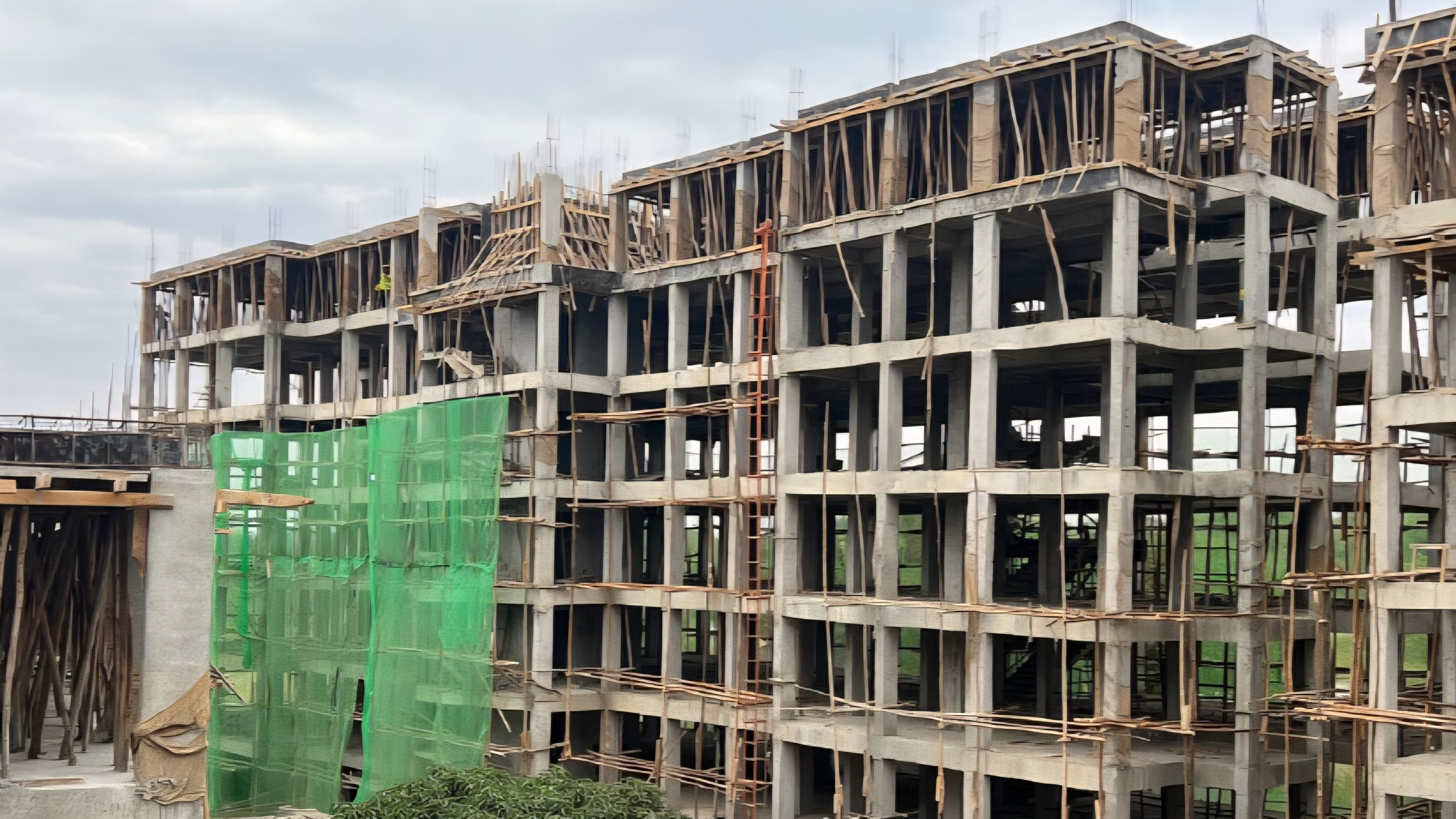 Concrete structural frame of a multi-storey residential building under construction with green safety netting and wooden scaffolding.