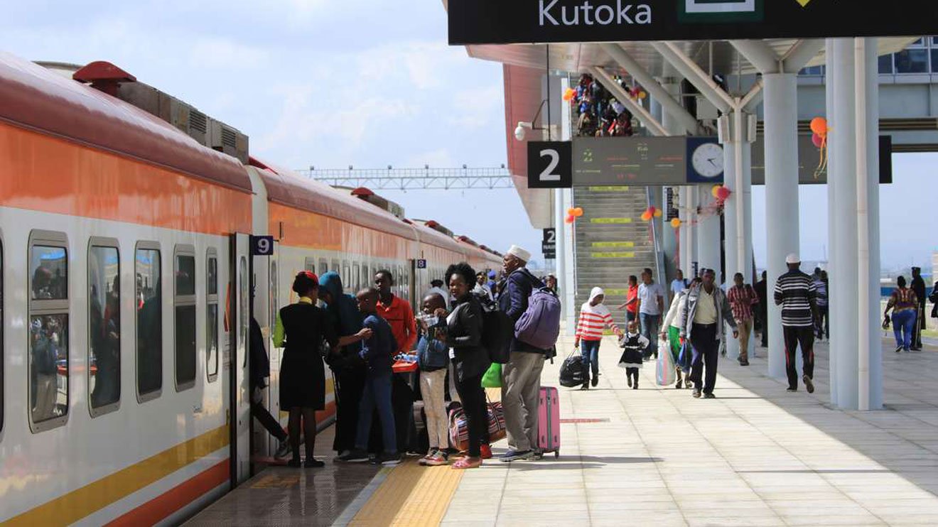 Passengers walking on the platform at the Nairobi SGR station with a passenger train and freight wagons visible on the tracks.