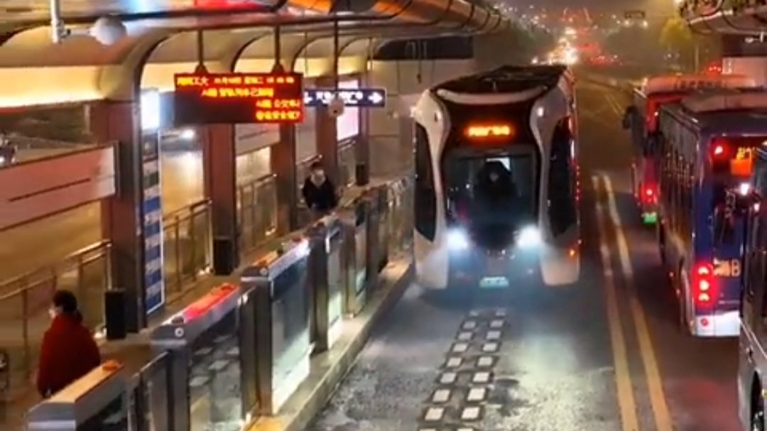 Nighttime view of white articulated tram approaching station on road with dotted virtual track lines and nearby buses.