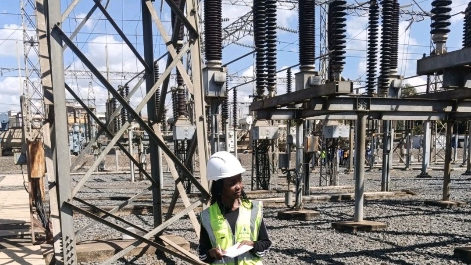 An engineer wearing a white hard hat and a high-visibility reflective vest stands in a gravel-covered electrical substation courtyard, surrounded by high-voltage steel towers and circuit breakers, as seen in file 254851.png.