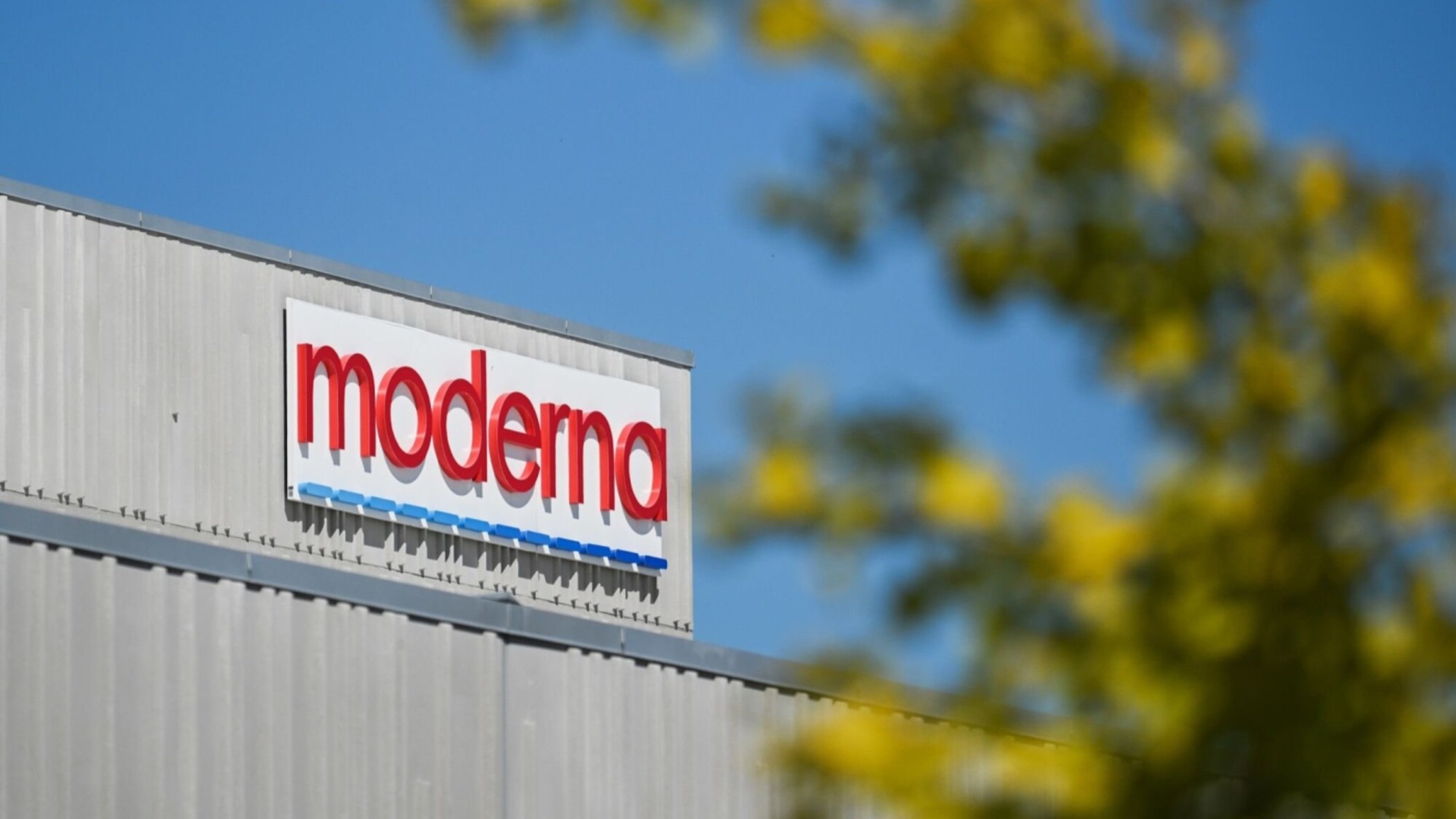 A exterior shot of a Moderna facility showing the company logo on a building wall under a blue sky.