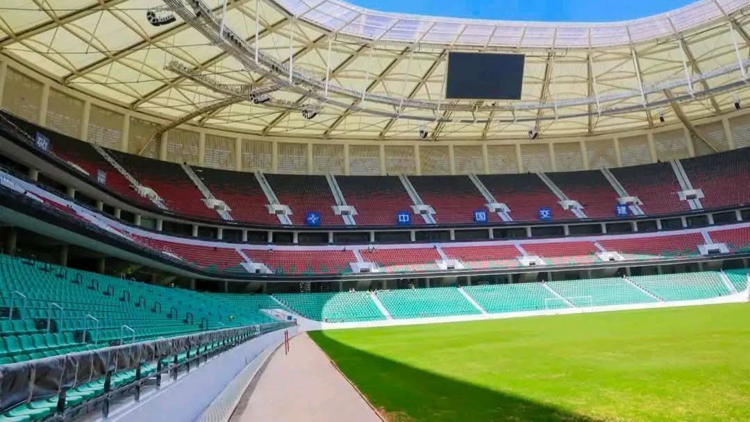 A view of a mini-stadium pitch with red seating stands, floodlights, and a grass field at the Talanta Sports City construction site.